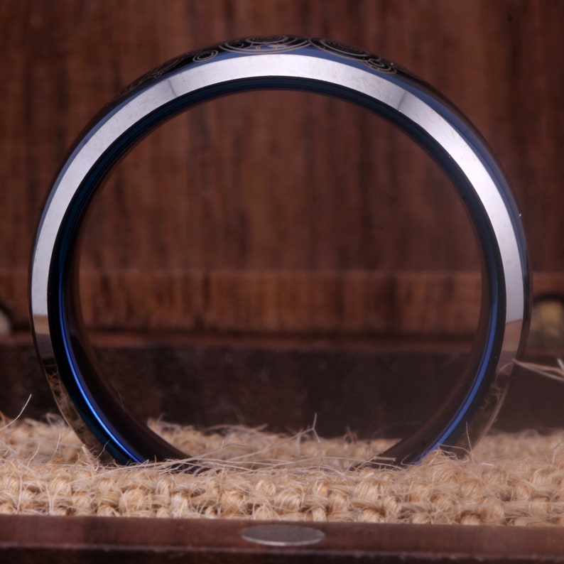 May include: A blue and silver tungsten carbide ring with a decorative design. The ring is shown from the side, with the inside of the ring facing the camera.