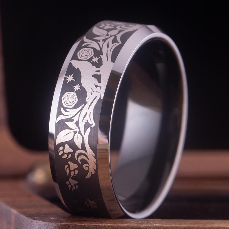 May include: A black tungsten carbide ring with a silver-colored design of a wolf howling at the moon, surrounded by leaves and paw prints.