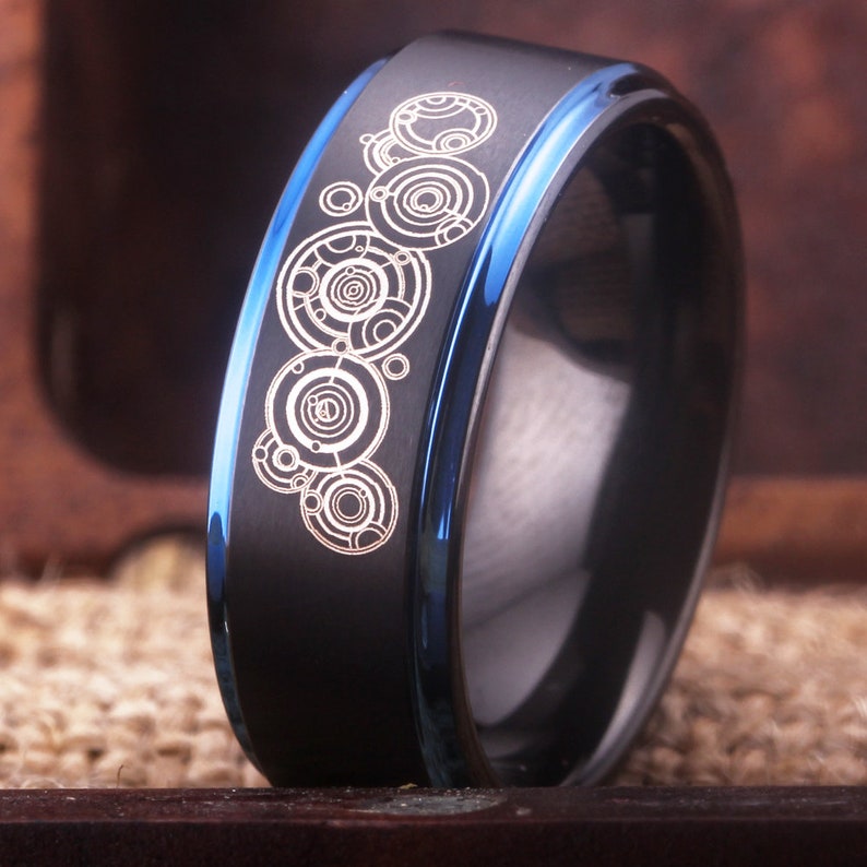 May include: A black and blue tungsten carbide ring with a silver design of a series of concentric circles.