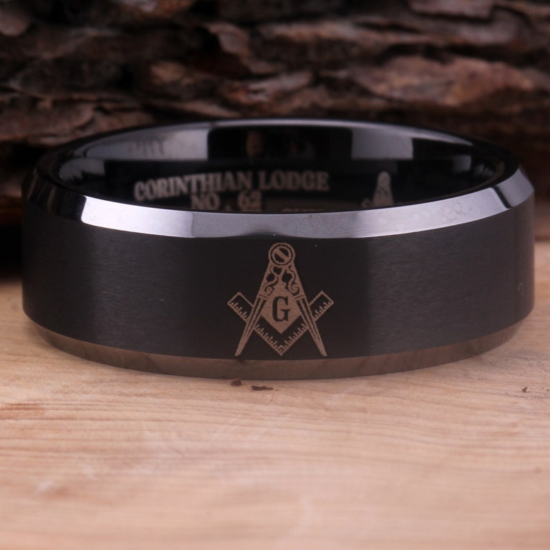 May include: A black tungsten carbide ring with a beveled edge and a laser engraved Masonic symbol and the text "Corinthian Lodge No. 62".