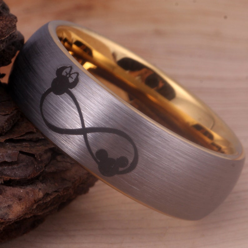May include: A silver tungsten carbide ring with a gold band and an infinity symbol with Mickey Mouse heads engraved on it.