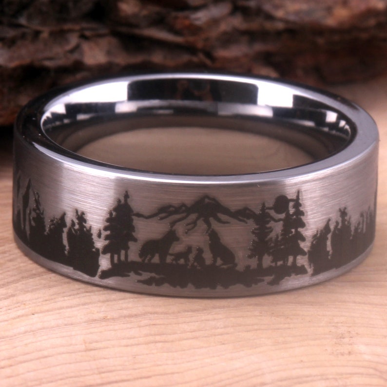 May include: A silver tungsten carbide ring with a brushed finish and a black engraved design of a mountain range, trees, and wolves.