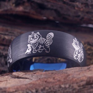 May include: Black ring with a blue interior and engraved white illustrations of the Cheshire Cat from Alice in Wonderland. The ring is set against a natural wood background.