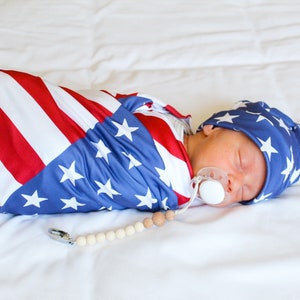 May include: A baby swaddled in a red, white, and blue American flag print swaddle. The baby is wearing a matching hat and has a pacifier in their mouth. A wooden bead necklace is on the white sheet.
