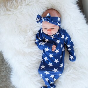 May include: A baby wearing a blue and white star patterned knotted gown and a matching headband. The baby is lying on a white fluffy surface.