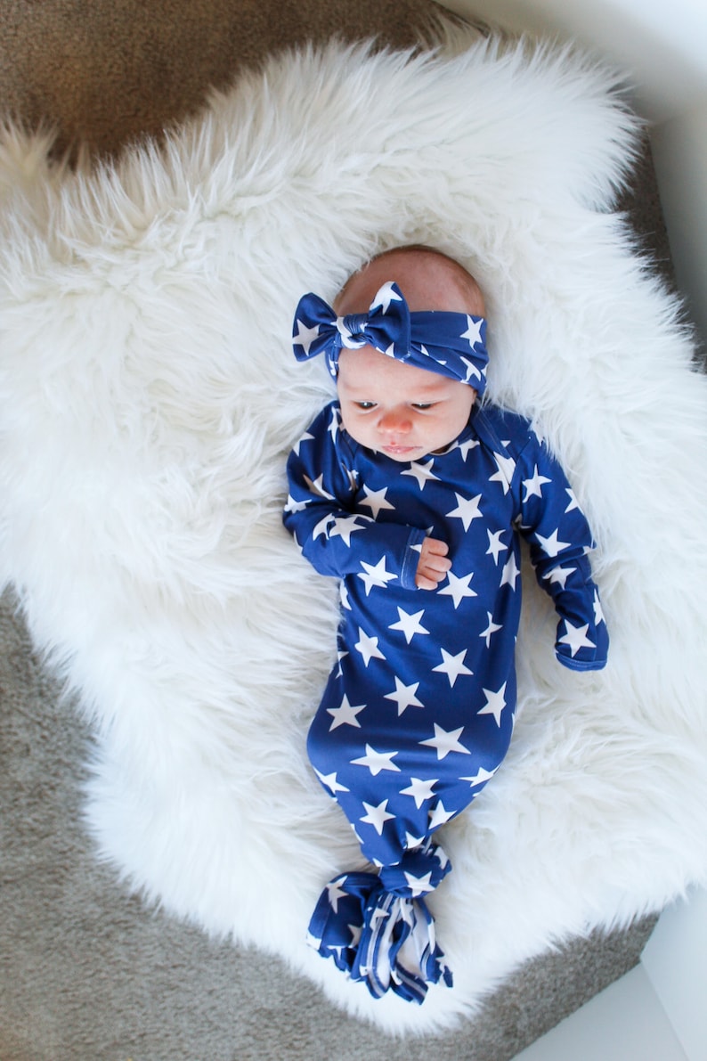 May include: A baby wearing a blue and white star patterned knotted gown and a matching headband. The baby is lying on a white fluffy surface.
