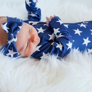 May include: A baby wearing a blue and white star patterned sleep sack with a matching blue and white star headband. The baby is lying on a white fluffy surface.