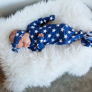 May include: A newborn baby wearing a blue and white star patterned knotted gown and matching headband. The baby is lying on a white fluffy rug.