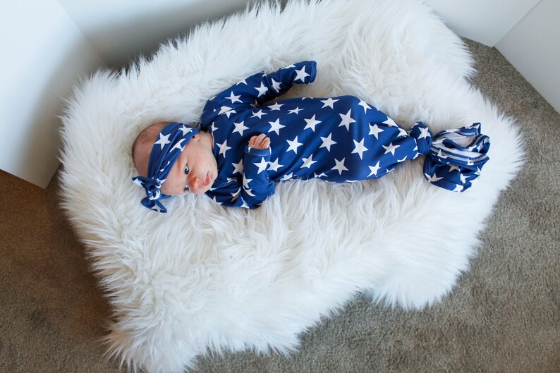 May include: A newborn baby wearing a blue and white star patterned knotted gown and matching headband. The baby is lying on a white fluffy rug.
