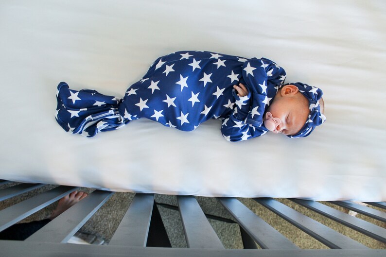 May include: A newborn baby sleeping in a blue and white star patterned swaddle. The baby is wearing a matching headband and has a pacifier in their mouth.