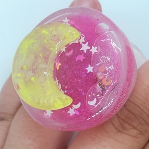 May include: A pink and clear resin sphere with a yellow crescent moon and white stars inside. The sphere is filled with glitter.