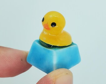May include: A yellow rubber ducky keycap with black eyes and an orange beak, sitting on a blue and white keycap.
