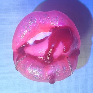 Puede incluir: Labios de resina rosa con un glaseado rojo que gotea y detalles de cadena plateada. Los labios están cubiertos de purpurina.