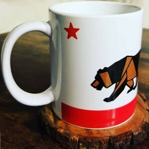 May include: White ceramic mug with a red star and a black bear silhouette on a red and white background. The mug is sitting on a wooden coaster.