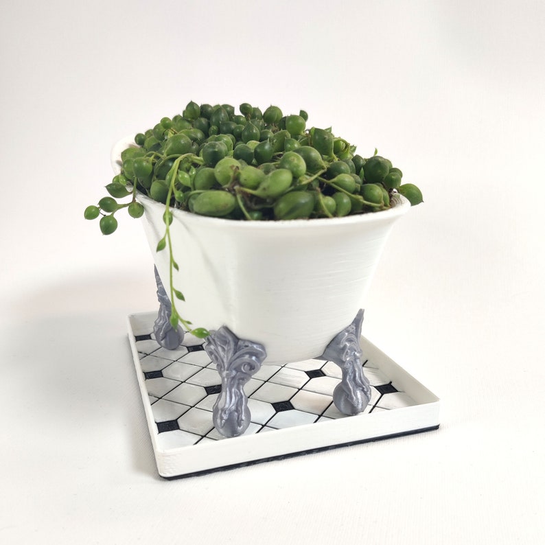 May include: A white ceramic bathtub planter with a green succulent plant. The planter has four decorative feet and is sitting on a black and white tiled tray.