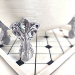 May include: A white bathtub with silver ornate feet. The bathtub is sitting on a black and white tiled floor.
