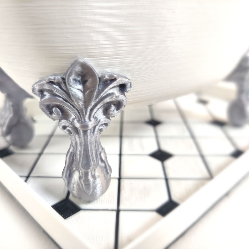 May include: A white bathtub with silver ornate feet. The bathtub is sitting on a black and white tiled floor.