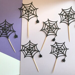 May include: Six black paper spider web cupcake toppers with small black spiders attached to the bottom. The toppers are on a purple and white background.