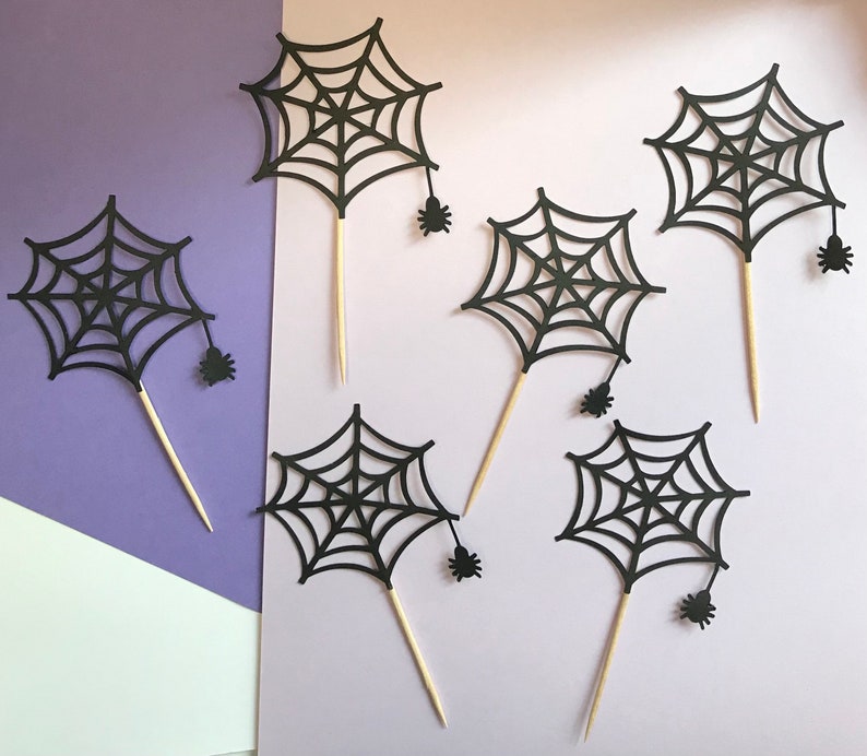 May include: Six black paper spider web cupcake toppers with small black spiders attached to the bottom. The toppers are on a purple and white background.