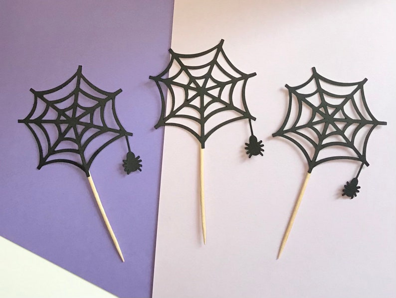 May include: Three black paper spiderweb cupcake toppers with small black spiders attached. The toppers are on a purple and white background.