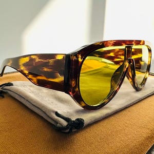 Może przedstawiać: Okulary przeciwsłoneczne w kolorze żółwim panterki z dużymi, okrągłymi, żółtymi szkłami. Okulary przeciwsłoneczne leżą na szarej materiałowej torbie.