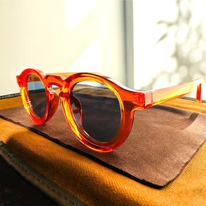 Op de afbeelding: Oranje getinte zonnebril met een rond montuur en donkere glazen. De zonnebril ligt op een bruin oppervlak. De monturen zijn doorschijnend oranje en de glazen zijn donker.