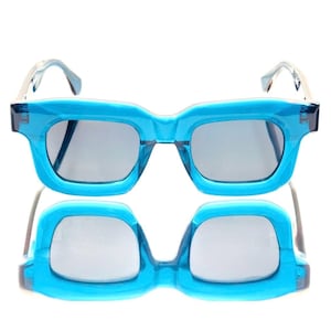 Puede incluir: Unas gafas de sol azul turquesa con una montura rectangular gruesa. Las lentes son grises y las patillas son blancas translúcidas. Las gafas de sol se reflejan debajo, creando una imagen simétrica.