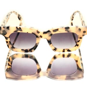May include: Sunglasses with a tortoiseshell design. The frames are a light beige with dark brown spots. The lenses have a gradient, going from dark grey at the top to a lighter grey at the bottom. The sunglasses are reflected below.