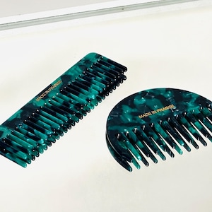 Könnte beinhalten: Zwei Haarkämme in einem marmorierten türkis-schwarzen Muster. Einer ist rechteckig, der andere hat eine halbkreisförmige Form. Beide Kämme haben Zähne und den Text "MADE IN FRANCE" in goldfarbenen Buchstaben.