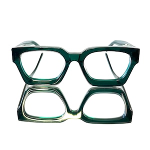 May include: A pair of emerald green eyeglasses with a thick, angular frame. The glasses have a square shape with rounded edges and clear lenses. The image shows the glasses reflected on a shiny surface.
