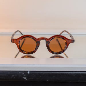May include: Round-framed sunglasses with a brown tortoiseshell pattern on the frame and temples. The lenses are a warm brown color. The glasses are displayed on a reflective surface.