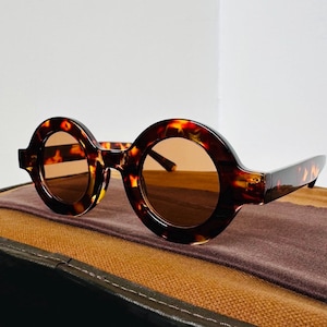 Puede incluir: Gafas de sol redondas de carey con lentes marrones. Las gafas de sol están sobre una superficie de cuero marrón.