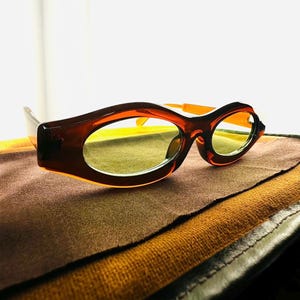 Puede incluir: Gafas de sol ovaladas marrones con lentes amarillas. Las monturas tienen un diseño grueso y redondeado. Las patillas son de color naranja brillante. Las gafas de sol están sobre una tela marrón.