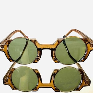 Puede incluir: Dos pares de gafas de sol de estilo vintage con lentes redondas verdes y una montura geométrica única en color ámbar translúcido. Las gafas tienen una estética retro, adecuadas para moda o disfraces.