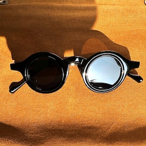 Puede incluir: Gafas de sol redondas negras con lentes oscuras. Las gafas tienen una montura negra gruesa y brillante y un revestimiento reflectante plateado en las lentes. Las gafas están sobre un fondo texturizado marrón anaranjado.