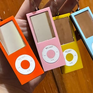 Puede incluir: Cuatro llaveros coloridos que son réplicas de un reproductor de música clásico en naranja, rosa, amarillo y azul. Cada uno tiene una pantalla y un panel de control circular con un botón de menú. Los llaveros son sostenidos por una mano.