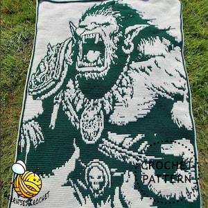 Puede incluir: Una manta de ganchillo verde y blanca que representa a un orco rugiendo. La manta tiene un diseño pixelado, con las palabras "CROCHET PATTERN" visibles. La manta se muestra sobre hierba.