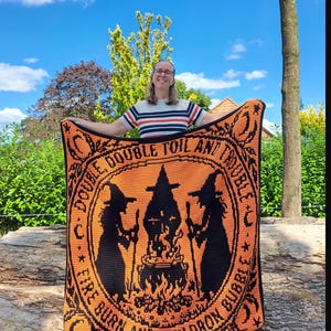 May include: A rectangular crochet blanket featuring a black and orange design of two witches standing on either side of a bubbling cauldron over a fire. The text "DOUBLE, DOUBLE TOIL AND TROUBLE, FIRE BURN AND CAULDRON BUBBLE" is printed in a circular border.