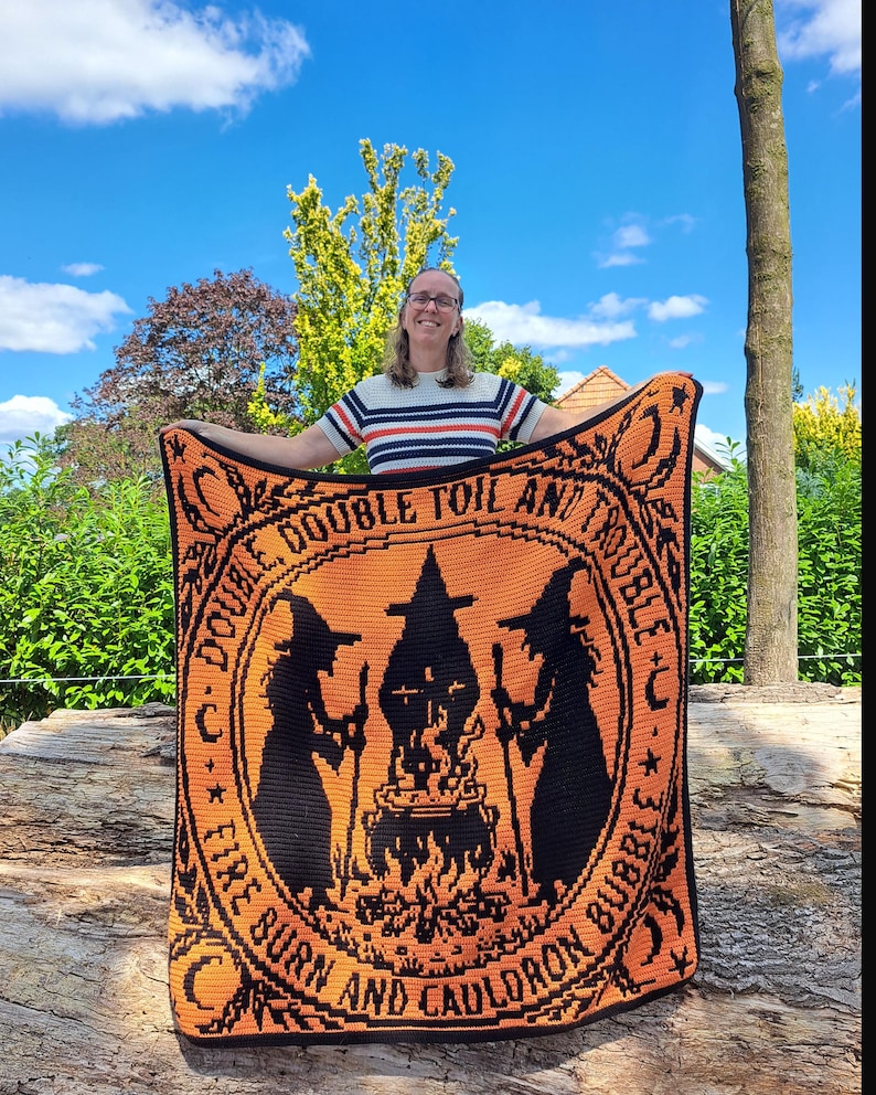 May include: A rectangular crochet blanket featuring a black and orange design of two witches standing on either side of a bubbling cauldron over a fire. The text "DOUBLE, DOUBLE TOIL AND TROUBLE, FIRE BURN AND CAULDRON BUBBLE" is printed in a circular border.