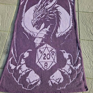 Puede incluir: Manta de ganchillo morada con un drag&oacute;n sosteniendo un dado de 20 caras. El drag&oacute;n es de color morado claro y el dado es de color morado oscuro.