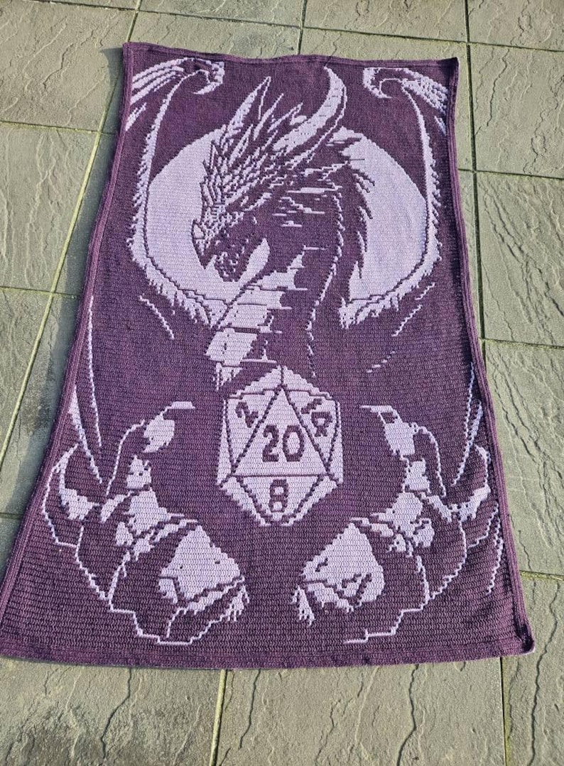 Puede incluir: Manta de ganchillo morada con un drag&oacute;n sosteniendo un dado de 20 caras. El drag&oacute;n es de color morado claro y el dado es de color morado oscuro.