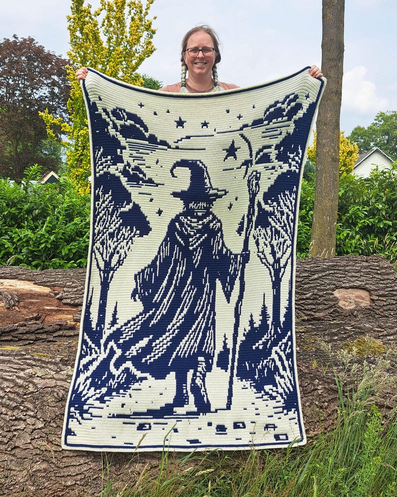 May include: A cream-colored crocheted blanket featuring a dark blue wizard design. The wizard is depicted with a staff, surrounded by trees and stars. The blanket has a dark blue border.