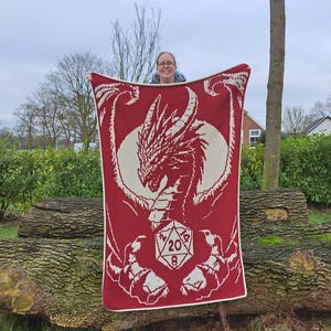 Puede incluir: Una manta de crochet roja con un dise&ntilde;o de drag&oacute;n blanco que sostiene un dado blanco de 20 caras. La manta est&aacute; siendo sostenida por una persona.