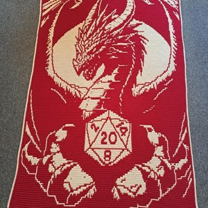Puede incluir: Una manta de crochet roja con un dise&ntilde;o de drag&oacute;n blanco que sostiene un dado blanco de 20 caras. Las alas del drag&oacute;n est&aacute;n extendidas y el dado est&aacute; en el centro de la manta.