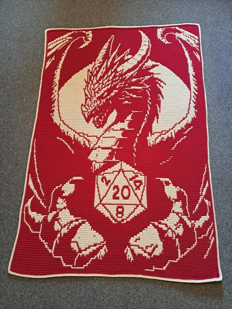 Puede incluir: Una manta de crochet roja con un dise&ntilde;o de drag&oacute;n blanco que sostiene un dado blanco de 20 caras. Las alas del drag&oacute;n est&aacute;n extendidas y el dado est&aacute; en el centro de la manta.