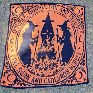 May include: A square crochet blanket featuring a black and orange design of two witches standing over a bubbling cauldron. The text "DOUBLE, DOUBLE TOIL AND TROUBLE, FIRE BURN AND CAULDRON BUBBLE" is written in a circular border with moon and star motifs.