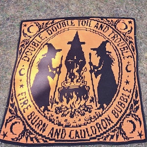 May include: A square crochet blanket featuring a black and orange design. The center depicts two silhouettes of witches standing by a bubbling cauldron over a fire. The text reads DOUBLE, DOUBLE TOIL AND TROUBLE, FIRE BURN AND CAULDRON BUBBLE.