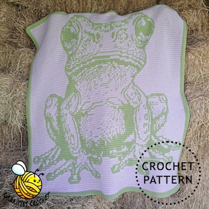 May include: A crocheted blanket featuring a large frog design in shades of green and lavender. The blanket has a green border and the words "CROCHET PATTERN" are in a circle on the lower right. The blanket is displayed on a bed of hay.
