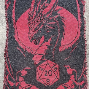 Puede incluir: Una manta de punto roja y negra con un dise&ntilde;o de drag&oacute;n que sostiene un dado de 20 caras.