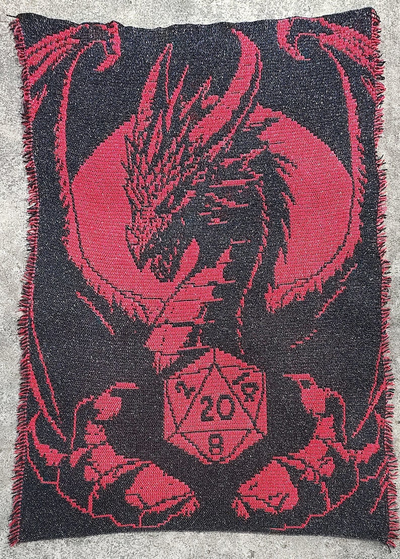 Puede incluir: Una manta de punto roja y negra con un dise&ntilde;o de drag&oacute;n que sostiene un dado de 20 caras.